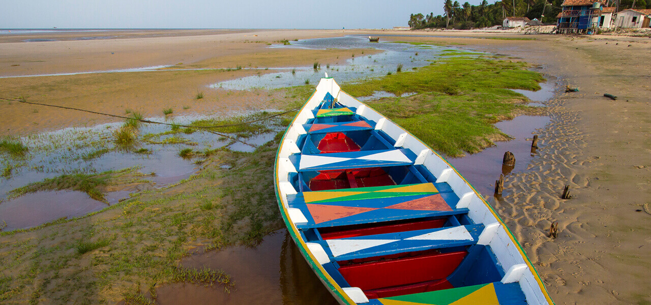Fotografia: Na praia, é dia Fotografia: sobre uma longa faixa de areia, uma canoa atracada com laterais internas azuis e bordas pintadas de branco. Os bancos são azuis e verdes, com pinturas triangulares brancas, vermelhas e amarelas. O piso é vermelho. A proa está voltada para o horizonte, sobre um banco de areia coberto por algas marinhas. À esquerda, ao fundo, o mar. À direita, algumas casas nas cores branca e azul, próximas a coqueiros.