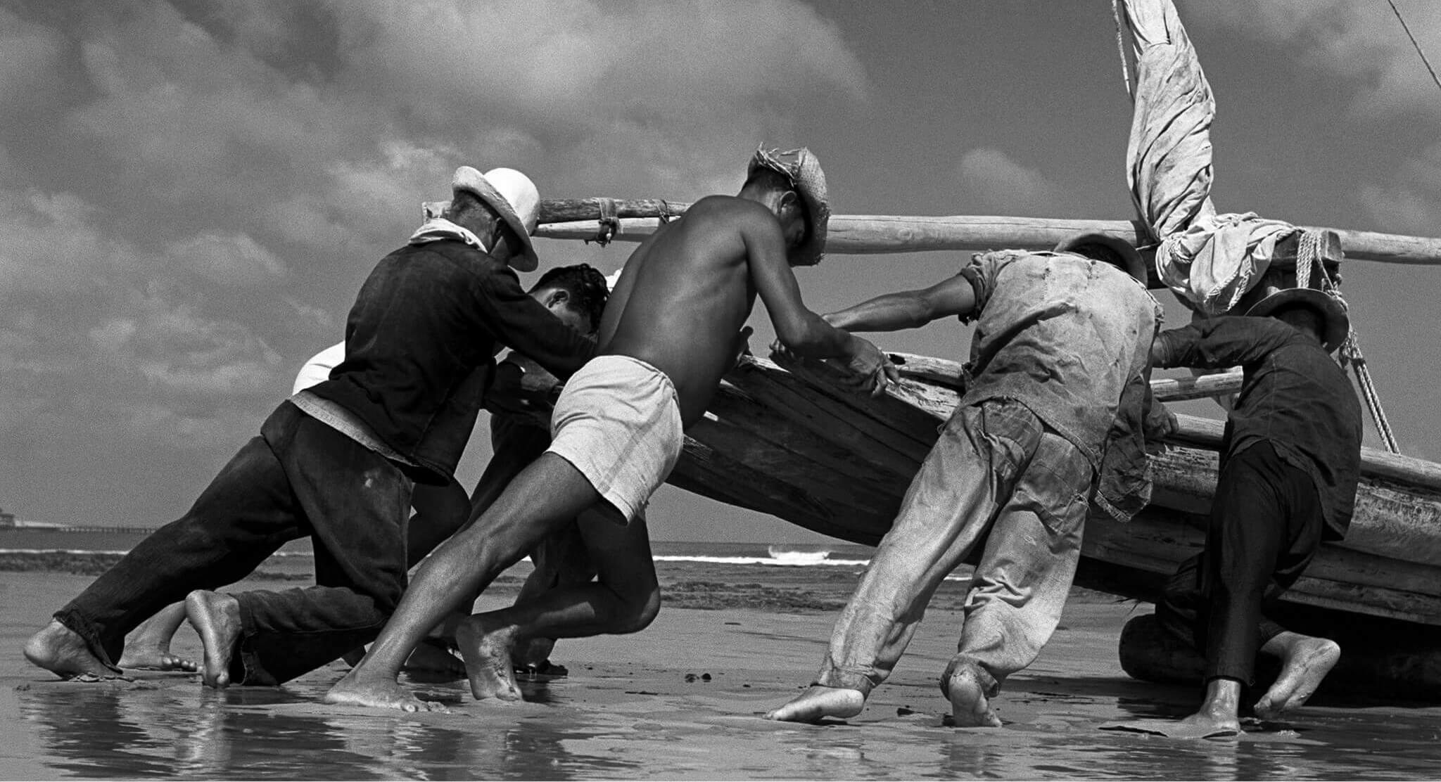 Jangadeiros Fotografia em preto e branco: Na praia, seis homens descalços, inclinam os corpos para frente e empurram um barco de madeira sobre a areia. A vela está enrolada e amarrada ao mastro. Ao fundo, o céu com nuvens e o mar.